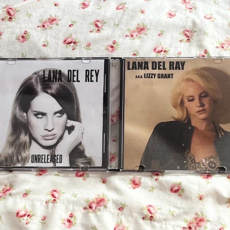 May include: Two CD cases featuring Lana Del Rey. One case has the text "Lana Del Rey" and "Unreleased" with a black and white photo. The other case says "Lana Del Rey a.k.a. Lizzy Grant" with a photo of the artist.