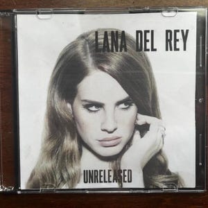 May include: A CD case featuring Lana Del Rey. The album cover is a black and white image of Lana Del Rey with the text "LANA DEL REY" at the top and "UNRELEASED" at the bottom. The image is in a clear plastic case.