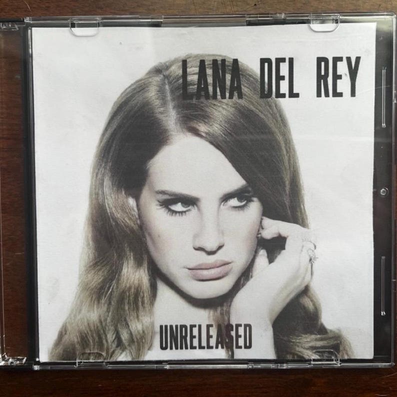 May include: A CD case featuring Lana Del Rey. The album cover is a black and white image of Lana Del Rey with the text "LANA DEL REY" at the top and "UNRELEASED" at the bottom. The image is in a clear plastic case.