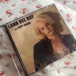 May include: CD case for Lana Del Rey's album, "Lana Del Ray a.k.a. Lizzy Grant." The album cover features a blonde woman in a black jacket and a sequined top. The CD case is clear, and the background is a light tan color.