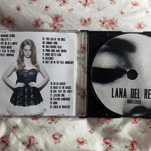 May include: A CD case with a photo of a woman in a black dress. The CD case is open, revealing the CD and tracklist. The CD is labeled "Lana Del Rey Unreleased."