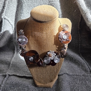 May include: A handcrafted necklace with a unique, artistic design. The necklace features brown, silver, and purple elements, including circular wooden pieces, wire spirals, and decorative metal accents. The necklace is displayed on a burlap bust.