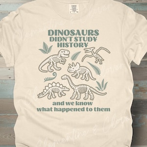 May include: A cream-colored short-sleeve t-shirt featuring a dark green graphic of four dinosaur skeletons and fern leaves. The text reads: DINOSAURS DIDN'T STUDY HISTORY and we know what happened to them. The shirt has a crew neck and a tag.