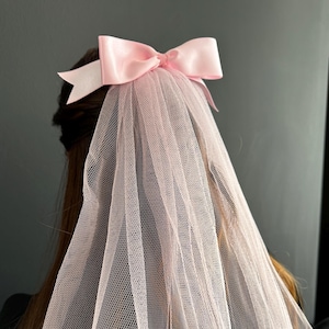 Puede incluir: Un velo de tul rosa claro con un lazo de satén a juego. El lazo está centrado en la parte superior del velo, que cae hacia abajo. Este delicado velo es un accesorio para bodas u ocasiones especiales.