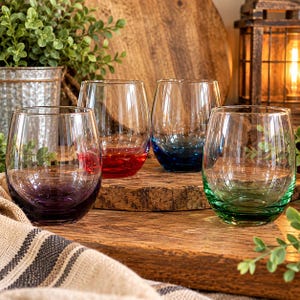 May include: A set of four stemless glasses with rounded bases, each featuring a distinct translucent color: purple, red, blue, and green. The glasses are clear at the top and transition into saturated, vibrant hues at the bottom, resting on a wooden surface.