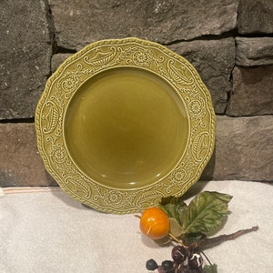Może przedstawiać: Zielony ceramiczny talerz z dekoracyjną kwiatową i przewijaną obwódką. Talerz ma falisty brzeg i gładki środek. Gałązka sztucznych jagód i mały pomarańczowy owoc znajdują się na pierwszym planie.