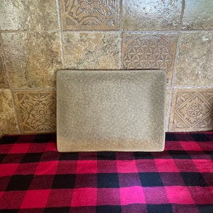 Puede incluir: Una bandeja rectangular de cerámica marrón claro con una superficie texturizada. La bandeja está sobre un mantel a cuadros rojo y negro.