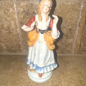 May include: A porcelain figurine of a woman wearing a blue hat, red and yellow dress, and white shoes. She is holding a basket in her right hand.