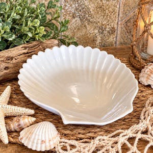 Op de afbeelding: Een witte keramische kom in de vorm van een grote sint-jakobsschelp met een geribbelde textuur en een gladde, glanzende afwerking. De kom is centraal geplaatst, omringd door een zeester, twee kleinere schelpen, een stuk drijfhout en een visnet.