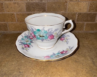 May include: A white teacup and saucer with a floral pattern in shades of pink, blue, and green. The cup and saucer have a gold trim.