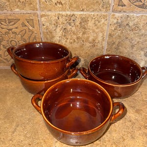 Puede incluir: Conjunto de cuatro cuencos de sopa de cerámica marrón con dos asas. Los cuencos están apilados uno encima del otro.
