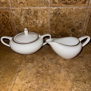 May include: A white ceramic sugar bowl and creamer set with a gold trim. The sugar bowl has a lid and the creamer has a spout.