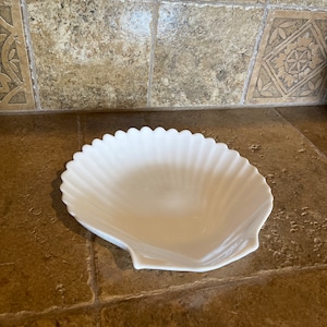 May include: A white ceramic seashell-shaped dish. The dish is smooth and has a fluted edge.