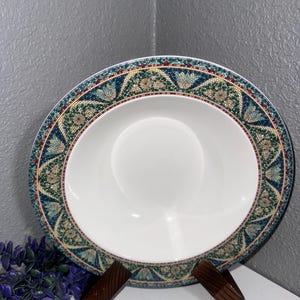 Könnte beinhalten: Eine weiße Keramikschüssel mit einem grünen, blauen und goldenen Mosaikdesign am Rand. Die Schüssel steht auf einem Holzständer.