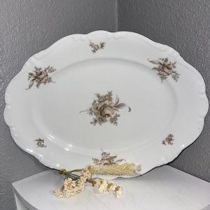 May include: White oval serving platter with a scalloped edge and floral rose pattern in shades of brown. The platter is displayed on a white shelf with dried flowers.