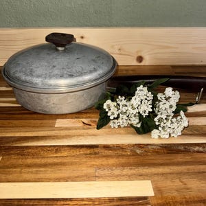 Puede incluir: Una olla de cocina de aluminio vintage con tapa y asa marrón oscuro. La olla es redonda con una tapa ligeramente abovedada y un pomo marrón oscuro. Un pequeño ramo de flores blancas con hojas verdes está cerca.