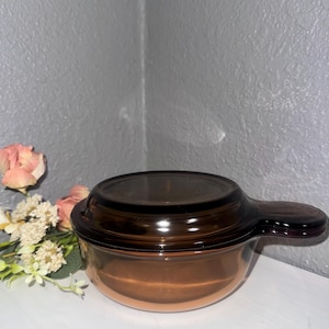 Op de afbeelding: Een bruine glazen ovenschaal met een deksel en een handvat. De schaal is rond en heeft een glad oppervlak.