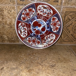 May include: A round ceramic plate with a gold rim and a colorful floral design. The plate features red, blue, and gold colors with a white background. The design includes stylized flowers, birds, and geometric patterns.
