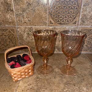 Pode incluir: Dois copos de vinho com haste transparentes de cor âmbar com um design em espiral. Uma pequena cesta de vime cheia de bagas pretas e vermelhas está sobre uma superfície de azulejos.