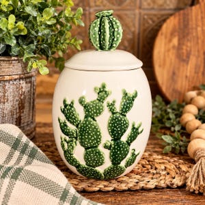 May include: A cream-colored ceramic canister with a rounded body and a matching lid topped with a small, green, textured cactus finial. The front of the canister features a raised, green prickly pear cactus design with white dots.