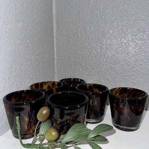May include: Set of six small, dark brown drinking glasses with a tortoiseshell pattern. The glasses are cylindrical with a slightly flared rim. A sprig of green leaves and olives is in the foreground.