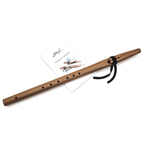 May include: A wooden flute with a dark finish, featuring a series of finger holes and a black ribbon detail. A white card with the text "Playing & Caring for" and a logo is placed near the flute.