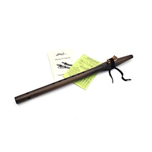 May include: A dark brown wooden flute with a smooth finish, featuring six finger holes along the body. A light-colored wooden block is secured to the top with a black leather cord. Two instructional pamphlets are positioned behind the flute.