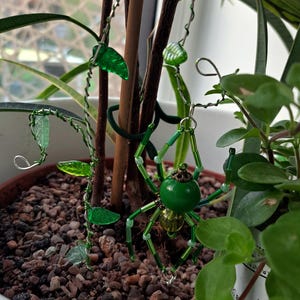 May include: A green beaded spider ornament with a round body and delicate legs, hanging amongst green glass leaf accents and a winding wire vine. The spider is near a potted plant.