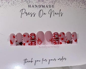 Könnte beinhalten: Handgemachte Press-on-Nägel mit einer rosa Basis und roten und weißen Designs. Die Nägel haben Herzen, ein Ringdesign und Strasssteine. Der Text "Setsbyserenity" ist auf der Karte sichtbar.
