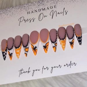 May include: Ten almond-shaped press-on nails featuring a matte mauve base with orange and white tips. The designs include black stitched patterns, orange pumpkin silhouettes, and orange and black animal print motifs on a display card with text.