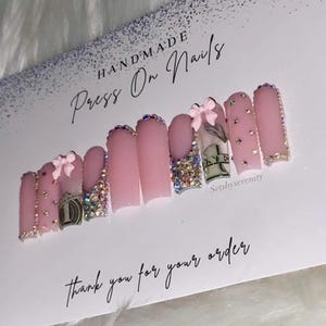 May include: Handmade press-on nails in a light pink hue. Each nail is adorned with rhinestones, bows, and dollar bill designs. The set includes a variety of embellishments, including glitter and gems. The card reads "Thank you for your order."