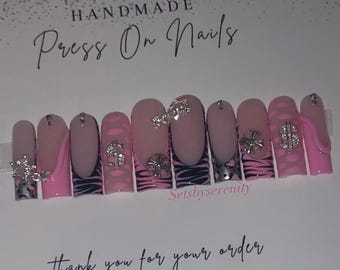 May include: Handmade press-on nails with a pink and black zebra print design. Each nail features a silver accent, including a dollar sign, a cross, and a butterfly. The nails are displayed on a white card with the text "Handmade Press On Nails" and "Thank you for your order".