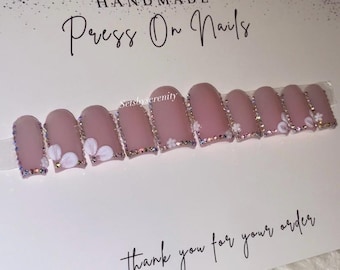 May include: A set of handmade press-on nails in a soft pink hue. Each nail is adorned with a delicate white floral design and a sparkling rhinestone border. The card reads "Thank you for your order."