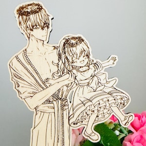 Peut inclure: Figurine en bois découpée au laser représentant une personne tenant un enfant, tous deux couronnés. La figurine est de couleur marron clair et repose sur une base en forme de rose. Un panier en céramique blanc rempli de roses roses est en arrière-plan.