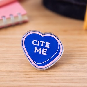 May include: A heart-shaped pin with the text "CITE ME" in white on a blue background, outlined in pink. The pin is on a wooden surface, with a pink notebook and pen in the background.