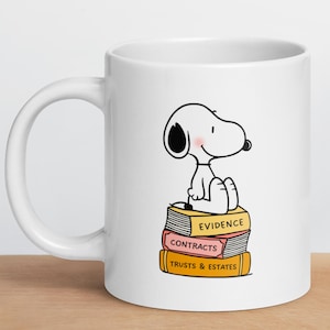 Puede incluir: Taza de cerámica blanca con un dibujo de Snoopy sentado sobre libros con las etiquetas "Evidence", "Contracts" y "Trusts & Estates". La taza tiene un asa curvada y un diseño sencillo.