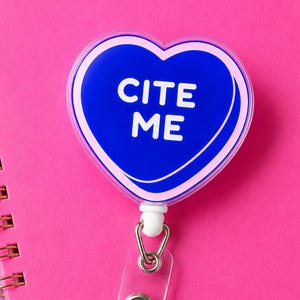 May include: A heart-shaped badge reel featuring a deep blue center with a light pink border. The text CITE ME is printed in white capital letters across the center of the heart. The badge reel includes a clear plastic strap and a metal snap button.