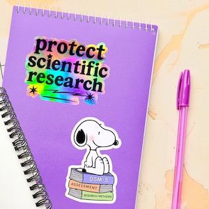May include: A purple spiral notebook featuring two stickers: one with the black text "protect scientific research" on a holographic background, and another showing a white cartoon dog sitting on a stack of books labeled DSM-5, ASSESSMENT, and RESEARCH METHODS.