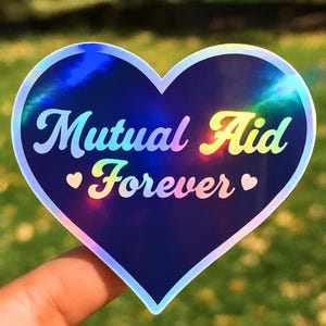 Op de afbeelding: Een hartvormige sticker met een holografische afwerking. De sticker bevat de woorden "Mutual Aid Forever" in een regenboogkleurige lettertype, met kleine hartsymbolen. De achtergrond is donkerblauw.