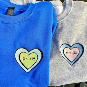 May include: Two sweatshirts, one blue and one gray, are displayed. Each has a heart-shaped patch with the text "p < .05". The blue sweatshirt has a light green heart, while the gray one has a pink heart.
