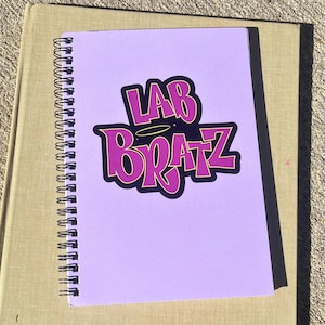 May include: A lilac spiral-bound notebook with the text "LAB BRATZ" in a purple, bubble-style font with a dark blue fill. The notebook is on a tan surface. The text has a gold outline.
