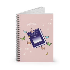 Puede incluir: Cuaderno de espiral rosa con la inscripci&oacute;n "psych girlie" y un gr&aacute;fico del libro DSM-5. La portada est&aacute; decorada con mariposas de colores y brillos. El cuaderno est&aacute; dise&ntilde;ado para los entusiastas de la salud mental.