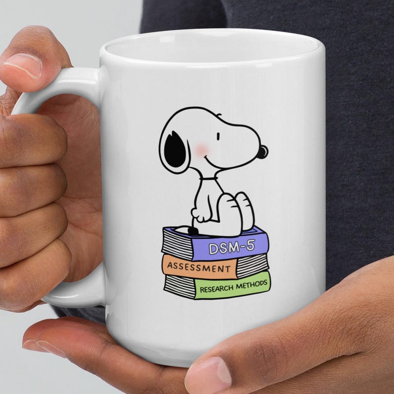 May include: White ceramic mug featuring a cartoon dog character sitting atop a stack of books labeled "DSM-5", "Assessment", and "Research Methods". The mug has a large handle and is held by a person.