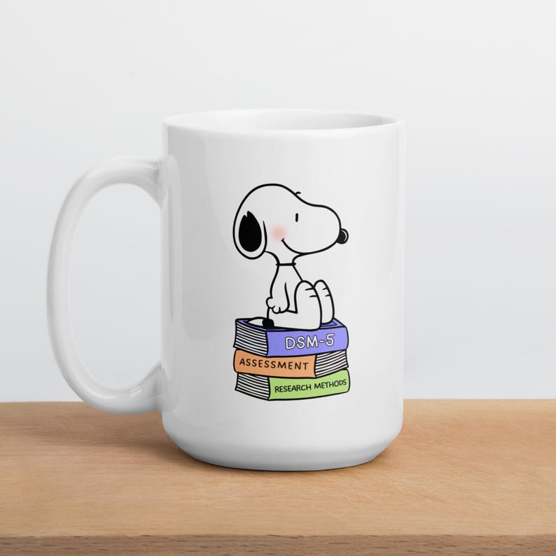 May include: White ceramic mug featuring a cartoon Snoopy character sitting atop a stack of books. The books are labeled "DSM-5", "ASSESSMENT", and "RESEARCH METHODS". The mug has a large handle and is set on a wooden surface.