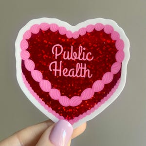 May include: A heart-shaped sticker with a red glitter center and pink frosting-like trim. The words "Public Health" are written in cursive script across the center. The sticker is held by a hand with a light pink nail polish.