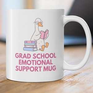 Peut inclure: Mug en céramique blanche avec un dessin d'oie lisant un livre rose, portant des lunettes et assise sur une pile de livres. Le mug porte l'inscription rose "GRAD SCHOOL EMOTIONAL SUPPORT MUG".