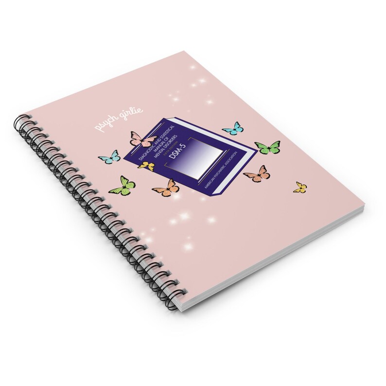 Puede incluir: Un cuaderno con espiral y tapa rosa claro. La portada presenta el texto "psych girlie" y un dise&ntilde;o que incluye un libro etiquetado como "DSM-5" y varias mariposas de colores. El cuaderno est&aacute; dise&ntilde;ado para tomar notas.