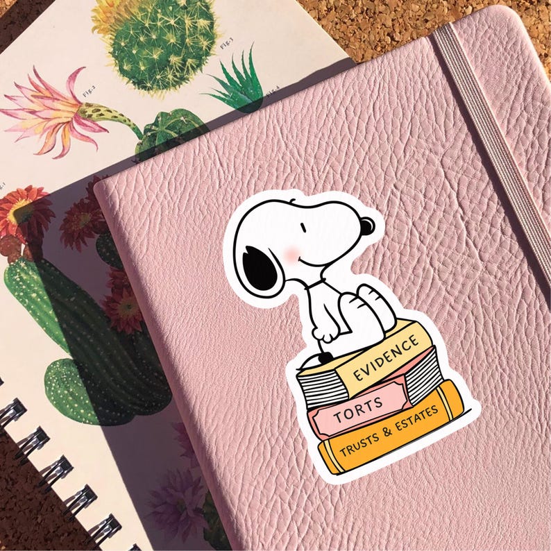 May include: A Snoopy sticker sits atop a stack of books labeled "Evidence," "Torts," and "Trusts & Estates." The sticker is white with black ears and a pink blush. The sticker is on a pink leather notebook.