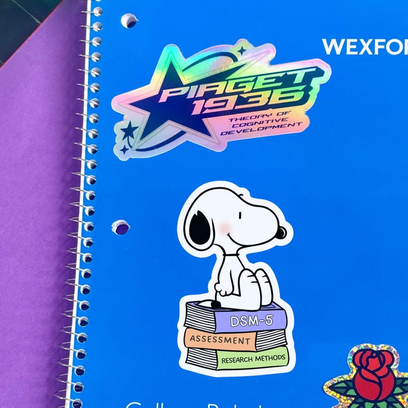 May include: A bright blue spiral-bound notebook decorated with three stickers. One sticker features a holographic star with the text Piaget 1936 Theory of Cognitive Development. Another shows a white dog sitting on a stack of books labeled DSM-5, Assessment, and Research Methods. A third sticker displays a red rose.