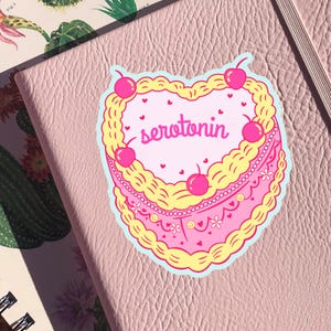 May include: A pink and yellow heart-shaped cake with white frosting and cherries. The cake says "serotonin" in pink letters.
